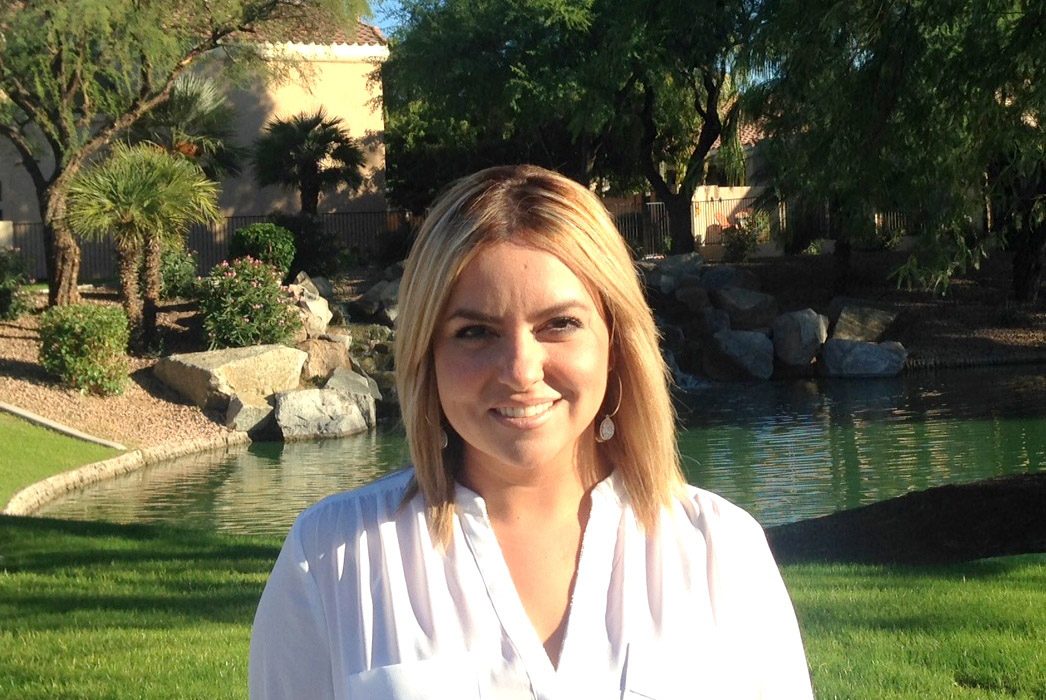 A woman smiles outdoors with greenery and a pond in the background.